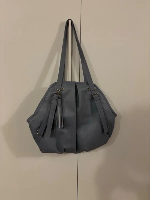 Blå axelväska med tofsdetalj - Snygg blå axelväska i mjukt material med långa axelband och dekorativ tofs framtill. Väskan har en modern, rundad form och metallringar som detaljer. Perfekt för dig som vill ha en stilren och rymlig väska till vardagen.