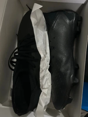 Adidas fotbollsskor svarta och Nike benskydd  - Säljer ett par svarta Adidas fotbollsskor med snörning och platt sula med dobbar för gräsplan. Skorna har en strömlinjeformad design och är tillverkade i syntetmaterial med textilinslag. Perfekt för dig som vill ha grymt grepp och lätt känsla på planen. Man får även benskydd med ett par Nike.