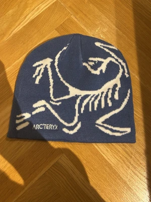 Blå mössa från Arc'teryx - Snygg blå mössa från Arc'teryx med vit grafisk design på framsidan och logotyp. Mössan är stickad och har en enkel, rundad form som sitter skönt på huvudet. Perfekt för kalla dagar och outdoor-aktiviteter.
