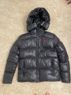 Svart pufferjacka från Polo Ralph Lauren - Svart pufferjacka från Polo Ralph Lauren med röd logga på bröstet. Jackan har huva med knappar, flera dragkedjefickor och quiltad design. Materialet är glansigt och känns riktigt robust, perfekt för kalla dagar. Snygg och stilren look med långärmade armar.