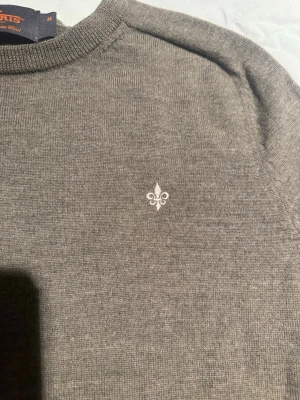 Grå stickad tröja från Morris - Snygg grå stickad tröja från Morris i 100% merinoull. Tröjan har rund hals, ribbade muddar och en liten broderad logga på bröstet. Perfekt för dig som gillar stilrena och klassiska plagg med en modern touch. Tröjan är lite liten i storlek så skulle säga att den passar S