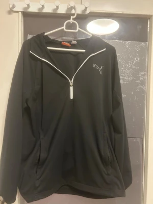 Svart träningströja  - Säljer en svart vindjacka från Puma med vit dragkedja i half zip-modell och huva. Jackan har diskret Puma-logga på bröstet och praktiska fickor på sidorna. Perfekt för dig som vill ha en sportig och clean look.