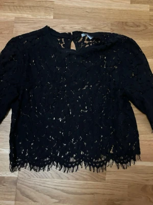 Svart spetsblus med lång ärm - Säljer en svart blus i spets med lång ärm och rund hals. Blusen har ett genombrutet mönster och fransad kant nedtill som ger en cool vibe. Perfekt att styla med jeans eller kjol för en trendig look.