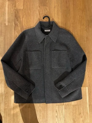 Marville Road Overshirt, strl 42 - Snygg mörkgrå overshirt från Marville Road med klassisk krage och två stora bröstfickor. Jackan är tillverkad i 100% mjuk ull och har knappar framtill samt vid ärmsluten. Något nopprig vid ärmslut och nedtill, men detta går att ta bort. Annars som ny! Storlek 42, men rätt liten i storleken. Jag har nu storlek 36 och den sitter som en oversized overshirt på mig- så beror lite på hur man vill att den ska sitta såklart :) Nypris: 2899kr och köpt på Åhléns city i Stockholm. Prislapp finns kvar.