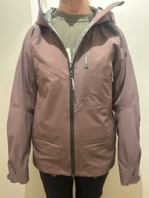 Lila Houdini skaljacka med huva - Snygg lila skaljacka från Houdini i storlek M. Jackan har huva, flera dragkedjeförsedda fickor och justerbara ärmslut. Materialet är poliester och jackan är perfekt för utomhusaktiviteter. Stilren design med diskret Houdini-logga på bröstet. Den har snöskydd längst ner på jackan. Passar perfekt på vandringen eller i skidbacken. Jackan kostar 7000 ny och är i väldigt bra skick utan defekter. 