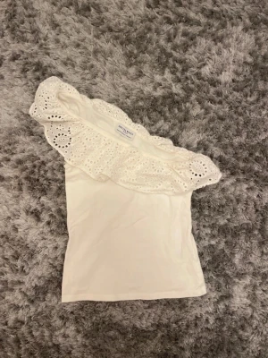 Vit oneshoulder topp från Holly & White - Säljer en vit oneshoulder topp från Holly & White med en bred volang upptill i broderad spets. Toppen är ärmlös och har en snygg asymmetrisk design med detaljerad spets som ger en romantisk vibe. Perfekt för varma dagar och sommarhäng.