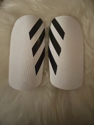 Adidas benskydd Tiro Shin Guards M - Säljer ett par Adidas Tiro Shin Guards i storlek M, perfekta för fotboll. De är vita med svarta ränder och har en snygg, sportig design. Tillverkade i syntetmaterial och kommer med en svart förvaringspåse med Adidas-logga. Passar dig som är mellan 140-160 cm.