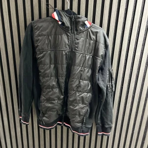 Svart quiltad pufferjacka från Moncler - Svart pufferjacka från Moncler med quiltad framsida och mjuka ärmar. Jackan har huva med röd, vit och blå kant, dragkedja framtill och ficka med dragkedja på ärmen. Ribbad mudd med röd, vit och blå detalj vid ärmslut och nederkant.