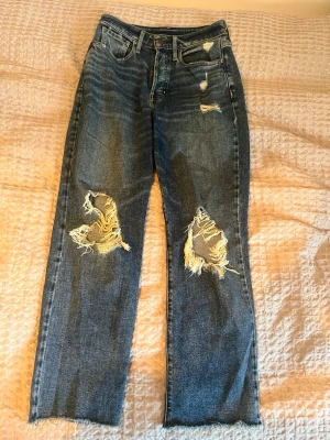 Blå ”boyfriend” jeans från Hollister - Säljer ett par blå jeans från Hollister med slitna detaljer och stora hål vid knäna. Modellen har raka ben, hög midja och knappar som gylf. Jeansen har fransad nederkant och klassisk femficksdesign. Perfekt för dig som gillar en edgy streetstil. Ingen lapp kvar, passar dock mig som har W26 / strl S.