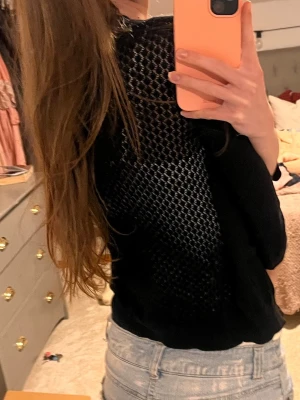Svart kofta med nätmönster - Säljer en svart kofta med snyggt nätmönster på ryggen och knappar framtill. Koftan har trekvartsärm och är gjord i ett tunt, stickat material som känns skönt mot huden. Perfekt att slänga över en topp för en chill och stilren look.