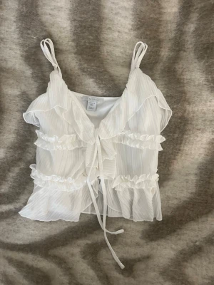 Vit volangblus från Amisu - Säljer en söt vit blus från Amisu i storlek XXS. Blusen har tunna axelband, volanger och knyt framtill. Materialet är lätt och transparent med diskreta ränder. Perfekt för dig som gillar romantisk och feminin stil.