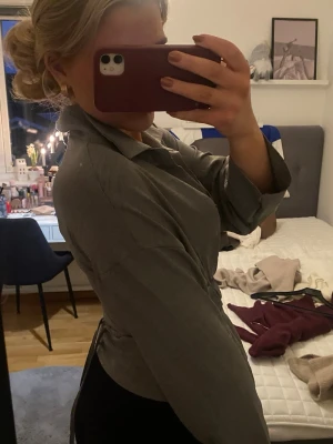 Grå skjorta med bälte och guldknappar - Snygg grå skjorta med markerad midja tack vare ett knytbälte. Skjortan har långa ärmar, klassisk krage och guldiga knappar framtill. Perfekt för dig som gillar en stilren och trendig look.