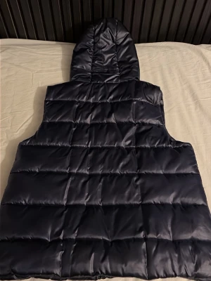 Mörk/Marinblå dunväst med huva från MILANO - Mörk/marinblå halvglossy dunväst från Milano med justerbar huva och dubbel dragkedja fram. Västen har knappar i sidorna för extra justering. Perfekt för att ha något under eller ovan och funkar året om. Västen har storlek 36 och är princip aldrig använd (Testad en gång). Jag säljer västen pågrund av att jag vuxit ur den. Nypriset var 1000kr och säljer den ny för 599kr, men priset är diskuterbart. 