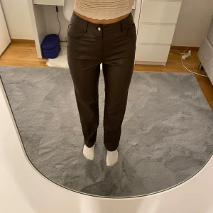 Bruna raka byxor i skinnimitation - Snygga bruna byxor i skinnimitation med raka ben och hög midja. De har klassisk femficksdesign, bälteshällor och knappstängning framtill. Materialet är slätt och ger en cool vibe till outfiten. Perfekt för dig som vill sticka ut lite extra.