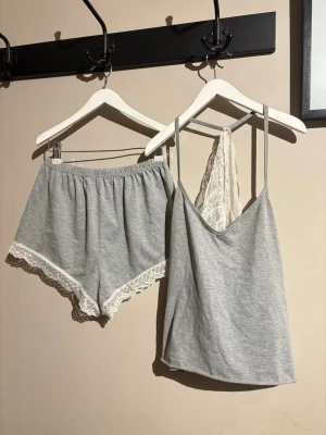 Grått set pyjamas - Säljer en ljusgrå linnetopp med smala axelband och vit spets i ryggen. Toppen är ärmlös och har en mjuk, flowy passform. Materialet är bomull med inslag av syntet, vilket gör den skön att ha på sig. Perfekt för chill dagar hemma eller som mysplagg.