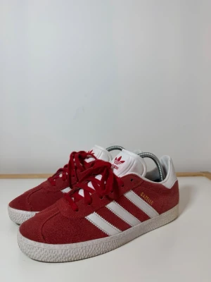 Adidas Gazelle röda sneakers - Säljer ett par Adidas Gazelle sneakers i rött mocka med vita detaljer och klassiska tre ränder på sidan. Skorna har vit plös med rött Adidas-tryck, snörning och vit sula. Perfekt för dig som gillar retrostil och vill sticka ut med färg.