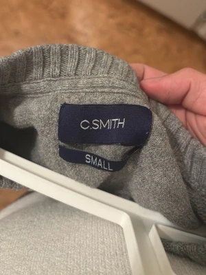 Grå stickad tröja C.SMITH Toronto - Grå stickad tröja från C.SMITH i storlek Small med broderad text 'SMITH TORONTO' och en kanadensisk flagga på bröstet. Tröjan har ribbade muddar vid ärmslut och nederkant samt rund hals. Perfekt för dig som gillar enkel och stilren design.