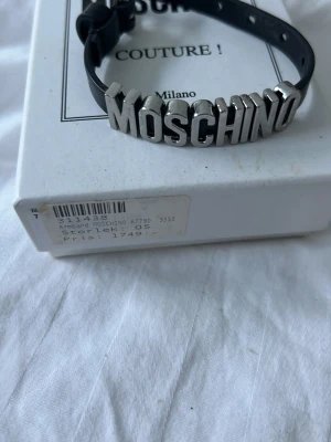 Svart och silver MOSCHINO armband - Coolt armband från Moschino med bred svart rem och stora silverfärgade bokstäver som stavar ut märket. Perfekt statement-accessoar för dig som gillar att sticka ut och visa din stil. Justerbar storlek och klassisk design.