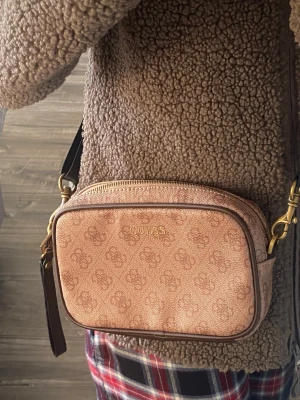 Brun axelväska från Guess - Modell: VEZZOLA I21 SMALL. Knappt använt. Kostade 75€ från början.  Väskan har justerbar svart axelrem, dragkedja upptill och flera fack inuti. 