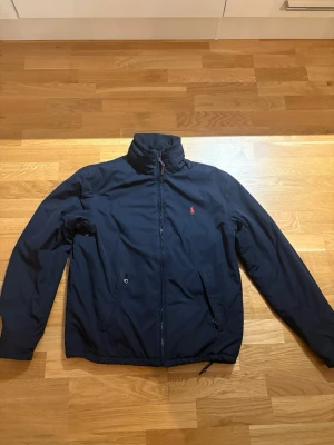 Vinterjacka Ralph Lauren - Snygg mörkblå vindjacka från Polo Ralph Lauren med röd logga på bröstet. Jackan har dragkedja, hög krage och två fickor framtill. Insidan är fodrad med mjuk fleece för extra komfort. Perfekt för kyliga dagar och enkel att matcha med din stil.