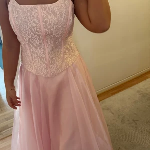 Ljusrosa långklänning med spetsliv - Drömmig ljusrosa långklänning med vackert spetsliv och markerad midja. Kjolen är i flera lager och ger en riktigt flowy känsla. Axelbandslös design med dragkedja bak. Perfekt för bal eller student!