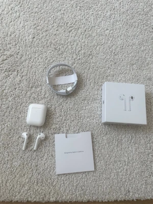Apple AirPods - Säljer ett par Apple AirPods med trådlöst laddningsetui, modell MRXJ2ZM/A. Komplett med originalkartong, laddkabel och manual. Hörlurarna är testade och fungerar, viss batterislitage syns på skärmbilden. Inga synliga repor eller skador på etui eller hörlurar.
