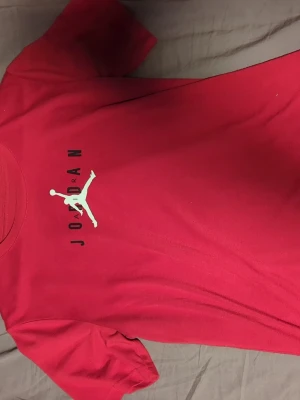 Röd Jordan t-shirt med logga - Säljer en röd t-shirt från Jordan med den klassiska Jumpman-loggan i vitt på bröstet och svart text. T-shirten har rund hals och korta ärmar, tillverkad i mjuk bomull som är skön mot huden. Perfekt för dig som gillar sportig streetstyle.