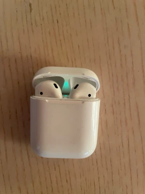 Apple AirPods (2nd generation) - Apple AirPods 2 med laddningsetui och originalförpackning. Trådlösa hörlurar med enkel anslutning till iPhone och andra Apple-enheter. Viss kosmetisk slitage syns på etuiet, men hörlurarna fungerar som de ska. Perfekt för musik, samtal och röststyrning.
