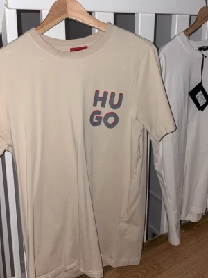 Beige t-shirt från Hugo - Säljer en beige t-shirt från Hugo med tryckt logga i rött och grått på bröstet. T-shirten har rund hals och korta ärmar, perfekt för en chill och stilren look. Materialet är mjuk bomull som känns skönt mot huden. I storlek L 