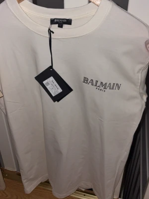 Vit t-shirt från Balmain Paris - Snygg vit t-shirt från Balmain Paris med klassisk rund hals och tryckt logga på bröstet. T-shirten är kortärmad och har en clean, minimalistisk look. Materialet är mjuk bomull som känns skönt mot huden. Perfekt för dig som gillar exklusiva märken och stilren design.