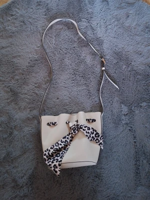 Beige axelväska med leoparddetalj - Snygg beige axelväska med justerbart axelband och coolt leopardmönstrat tyg som snörning och detalj framtill. Väskan har en bucket-form och är tillverkad i slätt syntetmaterial. Perfekt för dig som vill sticka ut med en trendig accessoar.