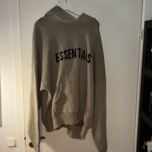 Beige Essentials hoodie stickad XL - Stickad hoodie från Fear of God Essentials i beige med svart logga på ryggen. Oversized fit, huva och känguruficka framtill. Perfekt för dig som gillar streetwear och vill ha en chill vibe.
