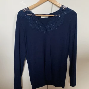 Mörkblå stickad tröja med spets och glitter - Mörkblå långärmad topp med v-ringning och dekorativ spets upptill, samt små glittriga detaljer längs halsringningen. Snygg och stilren modell som passar perfekt till jeans eller kjol. Vet tyvärr inte storlek men skulle gissa på att den är i storlek M då den är lite längre.