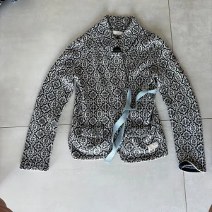 Mönstrad stickad cardigan jacka Odd Molly - Snygg stickad cardigan jacka från Odd Molly i blått och vitt med grafiskt mönster, ljusblå knytband i midjan och broderad kompassdetalj på ärmen. Jackan har lång ärm och är perfekt för lager-på-lager under höst och vinter.