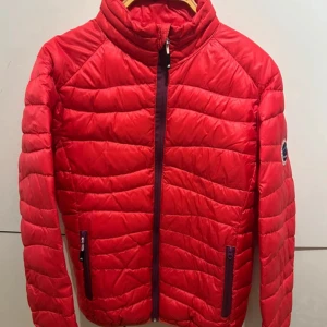 Röd pufferjacka från Tuxer, M - Riktigt snygg röd pufferjacka från Tuxer med quiltad design och hög krage. Jackan har dragkedja framtill, två fickor med dragkedja och ett coolt expeditionsmärke på ärmen. Perfekt för kalla dagar och outdoor vibes.