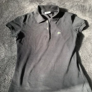 Svart Lacoste pikétröja i bomull - Svart pikétröja från Lacoste med klassisk krage och korta ärmar. Tröjan har två knappar framtill och den ikoniska gröna krokodilen broderad på bröstet. Materialet är mjuk bomull som andas och ger en skön passform.