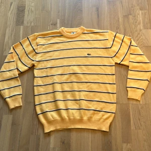 Gul randig stickad tröja från Lacoste - Gul stickad tröja från Lacoste med svarta och vita horisontella ränder. Klassisk rund halsringning och broderad krokodil-logga på bröstet. Tröjan har långa ärmar och ribbade muddar för en snygg passform. Perfekt för dig som gillar retrovibbar.