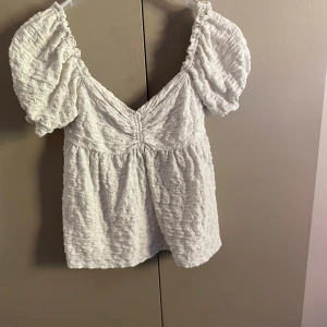 Vit blus med puffärm från Gina Tricot - Supersöt vit blus från Gina Tricot med puffiga korta ärmar och smockad struktur. Djup v-ringning framtill och snygg snörning i ryggen som ger en unik look. Perfekt för dig som gillar romantiska detaljer och vill sticka ut lite extra.