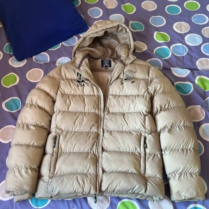 Beige pufferjacka Vinson Polo Club - Hej säljer en vinterjacka som heter polo club den är beige färg och den är väldigt bra att använda på vinter den är skönt och varmt och den har en stor fick in i jackan och sen har den två stora fickor och har använt den cirka tre år dock vill inte ha den längre hör av er om ni har några frågor.