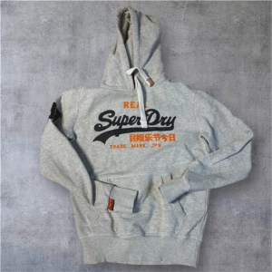 Superdry hoodie - Snygg grå hoodie från Superdry med stort svart och orange tryck på bröstet och japanska tecken. Tröjan har huva med snörning, magficka och ribbade muddar. Perfekt för chill dagar och streetstyle. Tillverkad i mjuk bomullsmix.