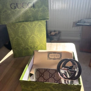 Beige bälte - Säljer ett bälte i beige och brunt med klassiskt monogrammönster och stor, rund metallspänne i silver. Bältet är gjort i skinn och har exklusiv känsla, perfekt för att lyfta vilken outfit som helst.