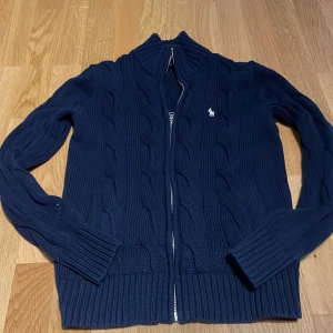 Mörkblå kabelstickad kofta Polo Ralph Lauren - Kabelstickad mörkblå kofta från Polo Ralph Lauren med dragkedja framtill och klassisk broderad logga på bröstet. Långärmad modell i mjuk bomull, perfekt för lager-på-lager. Snygg ribbad kant vid ärmslut och nederkant.