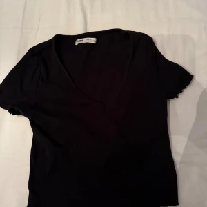 Svart v-ringad topp från Pull&Bear - En svart, enkel topp från Pull&Bear med v-ringning och korta ärmar. Toppen har lätt vågiga kanter vid ärmslut och nederkant, vilket ger en cool detalj. Perfekt att styla med jeans eller kjol för en avslappnad look.