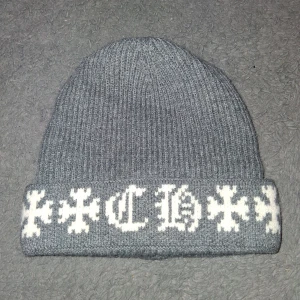 Grå Chrome hearts mössa  - Grå ribbstickad mössa med uppvikt kant och vit broderad text samt korsdetaljer i old english-stil runt kanten. Mjuk och varm, perfekt för kyliga dagar. Snygg streetwear-vibe och passar till de flesta outfits.
