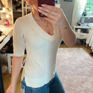 Vit v-ringad topp med kort ärm - Säljer en stilren vit topp med v-ringning och korta ärmar. Toppen har en mjuk och lätt ribbad känsla, slits i sidorna och sitter snyggt figurnära. Perfekt till jeans eller kjol för en clean look.