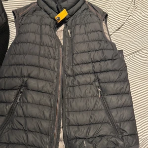 Svart dunväst från Parajumpers M - Svart quiltad dunväst från Parajumpers i superlätt material. Västen har dragkedja framtill, två fickor med dragkedja och en gul detalj med knapp vid kragen. Perfekt för lager-på-lager och snygg till streetwear. Finns ett litet hål