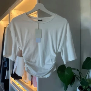 Vit croppad topp med snörning - Säljer en vit croppad topp från Soft Touch med korta ärmar och snygg snörning i sidan som ger en cool rynkad effekt. Toppen har rund halsringning och är gjord i mjuk bomull, perfekt för en trendig och avslappnad look.