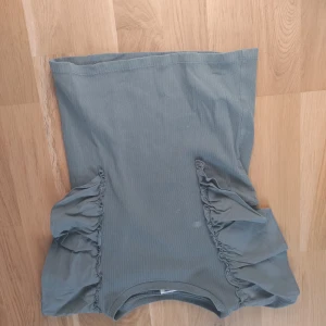 Grön ribbad tubtopp med volang från Kappahl - Snygg grön ribbad tubtopp från Kappahl i storlek 158/164. Toppen är ärmlös och har coola volangdetaljer på sidorna som ger en unik look. Perfekt för dig som gillar stilrena plagg med en twist.