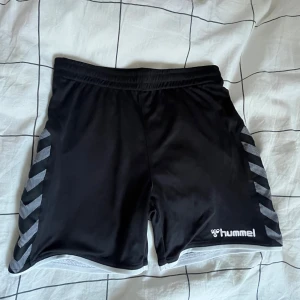 Svarta sportshorts från Hummel - Svarta sportiga shorts från Hummel med vita och grå ränder längs sidorna. Elastisk midja och hummel logga framtill. Perfekta för träning, säljer eftersom att dem är försmå, är inte använda så många gånger