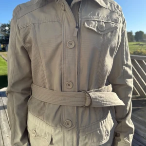 Beige jacka med bälte från Euro-Star - Snygg beige jacka från Euro-Star med markerad midja tack vare det breda bältet. Jackan har knappar, dragkedja och flera fickor framtill samt en cool krage. Tillverkad i bomull, perfekt för vår och höst. Långärmad och med stilrena detaljer.